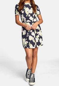 Robe bleu marine à manches courtes avec un grand motif floral aux couleurs pastel. Forme trapèze, tissu doux, longueur au-dessus du genou, associée à des baskets noires.