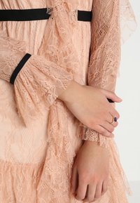 Händer korsade över persikofärgade spetsärmar med svarta detaljer, en hand bär en guldring med en mörk lila sten.