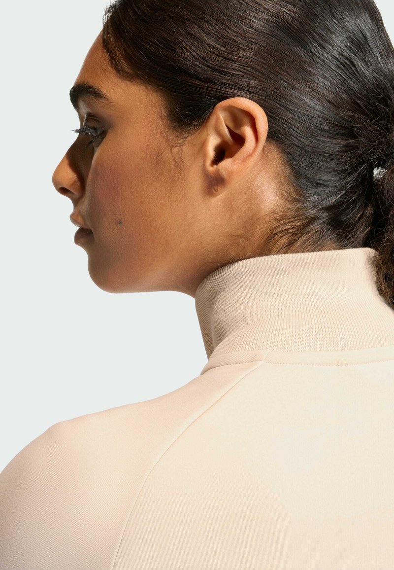 Top color beige chiaro con collo alto, realizzato in tessuto morbido ed elasticizzato. Presenta un design aderente e una texture liscia con cuciture sottili lungo le spalle.