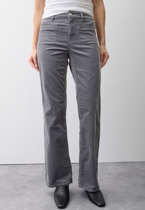Person wearing gray corduroy pants with front pockets, a white top, and black pointed-toe boots standing on a gray floor against a white wall.