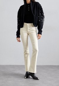 Cardigan noir en tricot avec des accents décoratifs, associé à un pantalon évasé crème et des bottines à bout pointu noires avec des embouts métalliques.