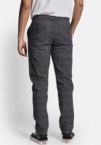 Grijze geruite broek met een gladde textuur, elastische tailleband en twee achterzakken. Gdraagd met zwarte sneakers met witte accenten.