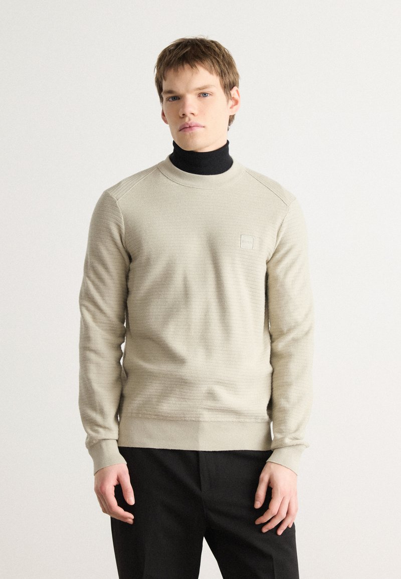 Beige tekstuuri sweatshirt, kus on ümmargune kaelus ja soonikkoes mansetid. Rinnal on väike logo plekk. Kantud musta kõrge kaelusega särgi peal.