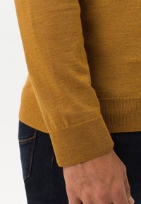 Pull jaune en maille avec poignets côtelés, coupe ajustée et texture lisse. Porté avec un jean en denim foncé.