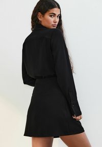 Abito camicia nero con maniche lunghe, colletto e vestibilità rilassata. Presenta dettagli con bottoni sui polsini e una texture del tessuto liscia.