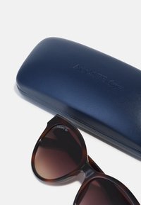 Slnečné okuliare s korytnačím vzorom a prechodovými hnedými šošovkami vedľa námornícky modrej, koženej puzdra s vyrazeným logom Lacoste.
