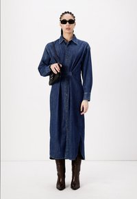 Robe chemise en denim avec taille ceinturée, manches longues, boutonnage à l'avant et fente sur le côté. Portée avec des lunettes de soleil noires et un petit sac noir.