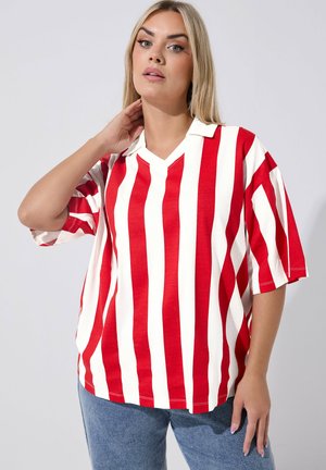 Donna bionda che indossa una maglietta a maniche corte a righe verticali rosse e bianche con colletto, abbinata a un paio di jeans blu, posando su uno sfondo bianco.