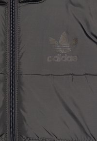 Šedá péřová bunda s hladkým, lesklým povrchem. Obsahuje centrální zip a vzorové logo Adidas ve tvaru trojlístku. Žádné viditelné vzory.