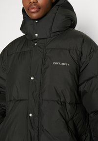 Zwarte pufferjack met hoge kraag, zilveren drukkers en een subtiel Carhartt-logo in het grijs geborduurd. Getextureerd quiltontwerp.