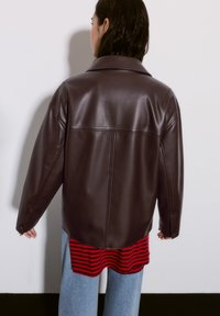 Persona che indossa una giacca di pelle marrone scuro sopra una maglietta a righe rosse e nere e jeans blu, di fronte a un muro bianco con un'ombra proiettata.