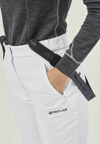 Weiße Hose mit schwarzen Akzenten und Logo, kombiniert mit einem dunklen strukturierten Oberteil. Verfügt über einen Reißverschluss und verstellbare Träger an der Taille.