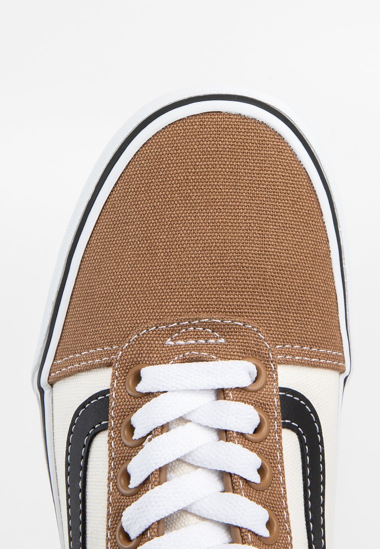 Zapatilla de lona marrón con cordones blancos, puntera crema, diseño de acento negro y suela de goma blanca. Superficie texturizada y detalles de costura visibles.