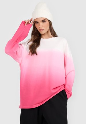 Kobieta w białej dzianinowej czapce i oversize ombre bluza przechodzącej z bieli w intensywny róż, w czarnych spodniach, pozuje na jednolitym tle.