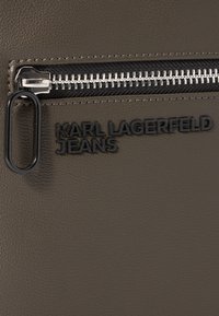 Tessuto sintetico grigio con finitura testurizzata. Presenta una tasca con zip e un logo in rilievo "KARL LAGERFELD JEANS" in nero.