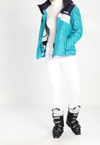 Turkusowa kurtka puchowa z czarnymi i białymi akcentami, nałożona na kremowy golf. Białe dopasowane spodnie i czarne buty narciarskie dopełniają wygląd.