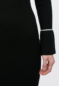 Zwarte geribde gebreide jurk met lange mouwen. De manchetten hebben een witte accentband. Close-up van de stoftextuur en het detail van de mouw.