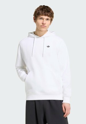 Młody mężczyzna w białej bluzie z kapturem Adidas z przednią kieszenią i czarnych spodniach, stojący na jasnym, jednolitym tle.