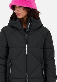 Giacca a vento nera con design a rombi trapuntati, chiusura con cerniera frontale, cappuccio e dettagli bianchi con logo. Il modello indossa un cappello in pelliccia artificiale di un vivido colore rosa.