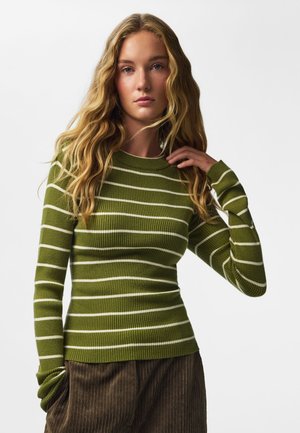 Ung kvinde med langt, bølget blondt hår iført en grøn- og hvidstribet ribstrikket sweater og brune fløjlsbukser.