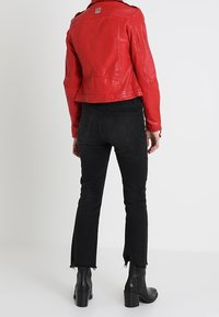 Vrouw die een geknipte rode leren jack, zwarte hoog-waist jeans met rauwe randen en zwarte hakken enkellaarsjes draagt, met haar rug naar de camera gekeerd.