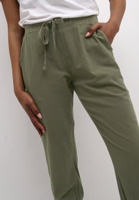 Personne portant un pantalon vert olive à cordon de serrage avec une taille élastique, les mains posées, une dans la poche, sur un fond uni.