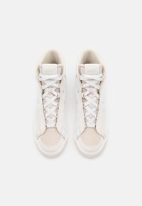 Korkeat tennarit, joissa on kermaisuus nahkamateriaali, valkoiset nauhat ja harmaat yksityiskohdat. Sileä pinta, pyöreä kärki ja huomattava logo kielellä.