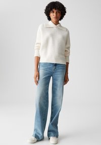 Maglione a coste di colore crema con colletto, abbinato a jeans a campana di un blu chiaro. Il modello indossa sneakers bianche. Lo sfondo semplice mette in risalto i dettagli dell'outfit.
