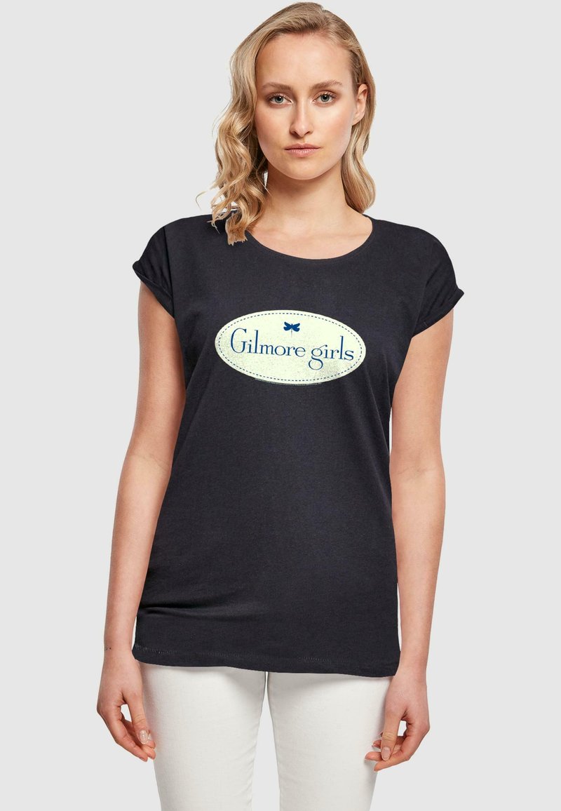 Juodas medvilninis marškinėliai su suktomis trumpomis rankovėmis, turintys didelį, ovalų, šviesiai žalią grafiką su užrašu "Gilmore girls" mėlyna spalva.