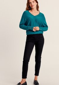 Pull v-neck teal à manches longues avec des détails de boutons argentés aux poignets, associé à un jean skinny noir et des mocassins noirs.