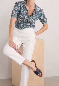 Camicia a maniche corte con stampa floreale in teal, abbinata a pantaloni bianchi con risvolto e sandali in tessuto blu navy; tiene occhiali da sole rosa.