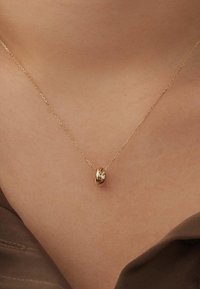 Collier en chaîne dorée avec un pendentif lisse et arrondi. La chaîne a une texture fine et est délicatement maillée, reposant au-dessus de la clavicule.
