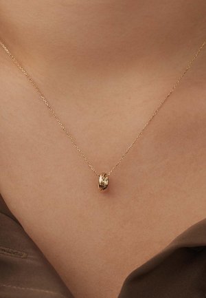 Collier en chaîne dorée avec un pendentif lisse et arrondi. La chaîne a une texture fine et est délicatement maillée, reposant au-dessus de la clavicule.