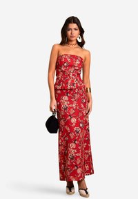 Rode bloemenmaxi-jurk met strapless peplumtop, aansluitende rok en ingewikkelde bloemmotieven. Gepaard met een zwarte handtas en espadrilles.