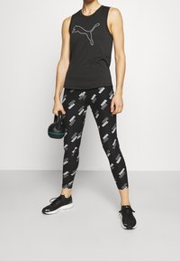 Top negro sin mangas con logo blanco, combinado con leggins negros que presentan un patrón gráfico blanco repetido. Modelo sosteniendo una kettlebell.