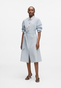 Ανοιχτό μπεζ πουκάμισο φόρεμα με διπλωμένα μανίκια, ζώνη στη μέση και μπροστινή σχίση. Διαθέτει χρυσά κουμπιά και λεία υφή υφάσματος.