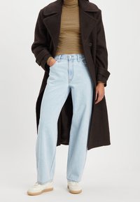 Lång brun kappa, randig polotröja, ljusblå vida jeans och vita sneakers med bruna sulor. Egenskaper inkluderar stor krage och detaljer med knappar.