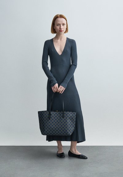 Robe côtelée gris foncé avec un profond décolleté en V et des manches longues. Ballerines noires et sac fourre-tout à motifs avec des accents bleus et dorés. Décor minimaliste.