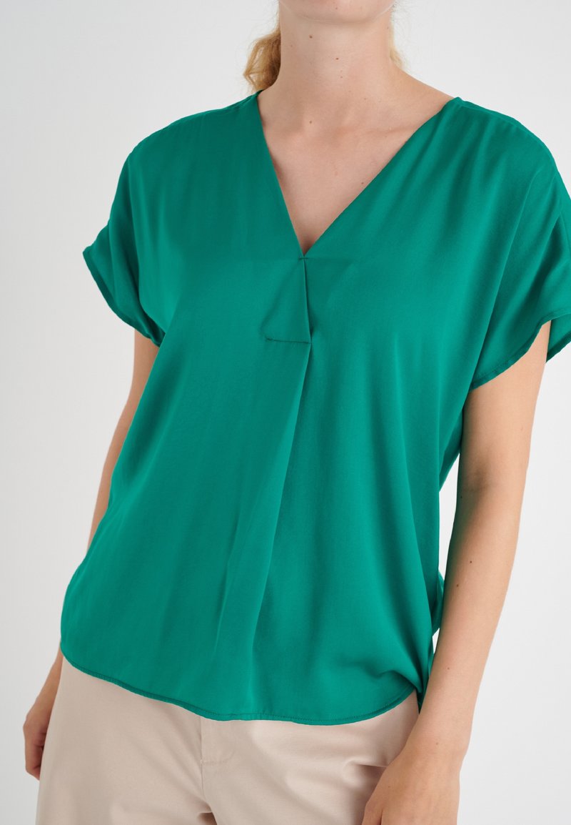 Femme portant un chemisier vert ample à manches courtes avec un col en V et un pantalon beige, photographiée du cou aux hanches.