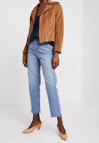 Veste de motard en daim marron avec des zips argentés, un top à motifs navy, un jean court bleu clair et des mules à talons beiges.