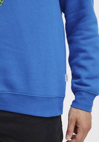 Felpa blu in misto cotone con polsini e orlo a coste. Presenta un piccolo logo laterale. Texture liscia, maniche lunghe.