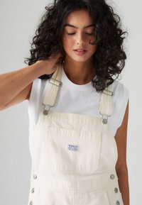 Salopette en jean couleur crème avec bretelles réglables, quincaillerie en métal, poches plaquées et étiquette logo sur la poitrine. Portée par-dessus un t-shirt blanc.