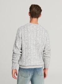 Szary dziergany sweter o prążkowanej fakturze i wzorach warkoczy, z okrągłym dekoltem i długimi rękawami. Połączony z jasnoniebieskimi dżinsami.