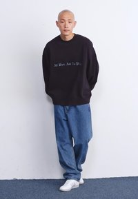 Felpa nera con la scritta "Tutto lavoro e niente svago" in un font chiaro, abbinata a jeans di denim blu larghi e sneaker bianche.