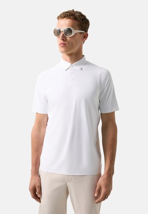 Man draagt een witte poloshirt met korte mouwen, beige broek en reflecterende ronde zonnebril, staand tegen een effen lichte achtergrond.