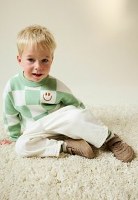 Junger blonder Junge sitzt auf einem cremefarbenen Shag-Teppich, trägt einen grün-weiß karierten Pullover mit einem Smiley-Gesicht und weiße Hosen.
