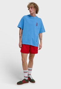 T-shirt oversize bleu clair avec un petit motif, assortie à un shorts rouge, des chaussettes blanches avec des accents rayés et des baskets en daim vert.