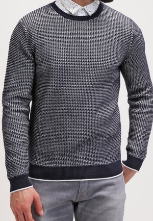 Mand iført en marineblå og hvid tekstureret striktrøje over en hvid skjorte med sorte pletter og lysegrå jeans.