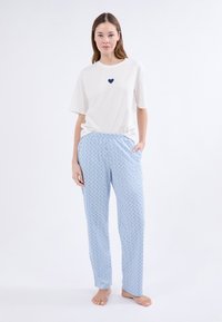 Mujer con una camiseta blanca de manga corta con un pequeño corazón en el pecho y pantalones de pijama ligeros de color azul claro con estampado, de pie descalza.