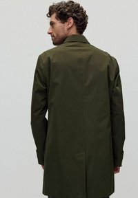 Oliivivihreä takki, jossa on muotonsa mukautettu malli, sileä rakenne, perinteinen kaulus ja napilliset hihansuut; takaa ulottuu puoleen reiteen.